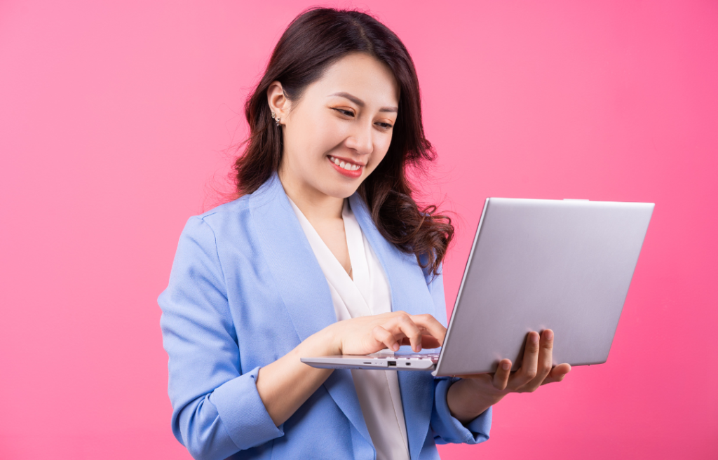 Smiling woman in a light blue blazer using a silver laptop against a pink background.