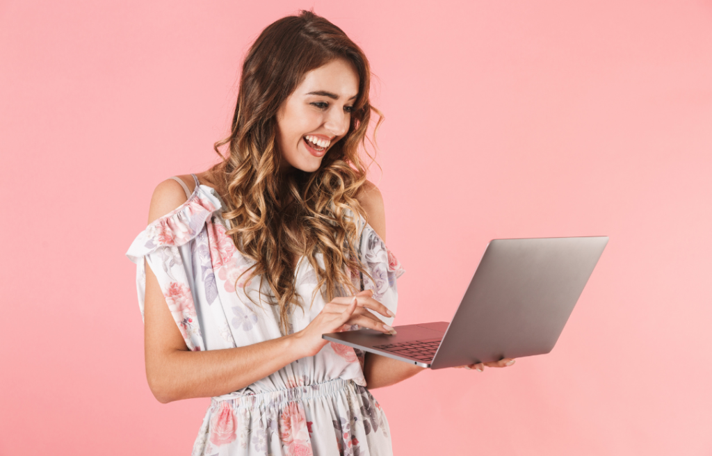 Woman in a floral dress using a laptop, smiling broadly against a pink background.