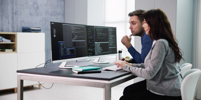 Two people working together at a desk with two computer monitors showing code.